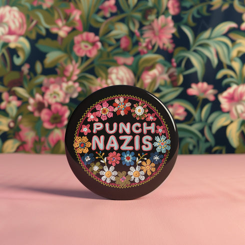 Round pinback metal button featuring an embroidered-style design on a black background. The center showcases bold, stitched-textured lettering in red, blue, and white that reads "PUNCH NAZIS." Surrounding the text is an intricate, folk-inspired floral pattern with multicolored flowers in shades of pink, orange, blue, and white, arranged symmetrically. A decorative zigzag border frames the edge of the button. The backside is silver metal with a secure pin closure.